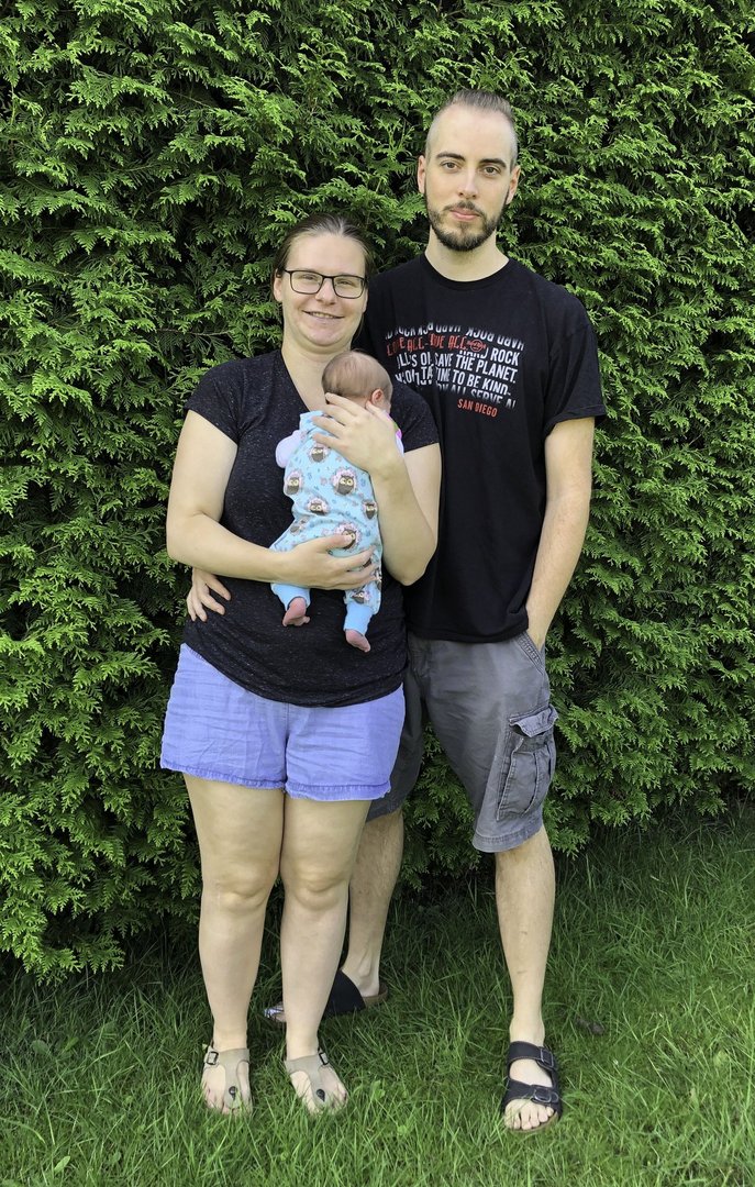 Frau und Mann mit Baby stehen vor einer grünen Hecke