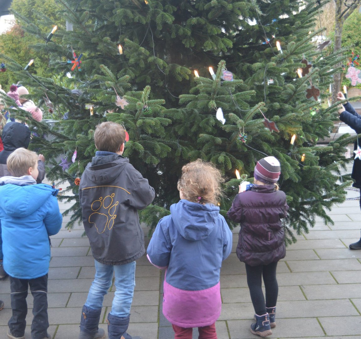 Kinder stehen vor einem großen geschmückten Weihnachtsbaum und betrachten selbst gebastelten Baumschmuck im Außenbereich des AKH Celle.