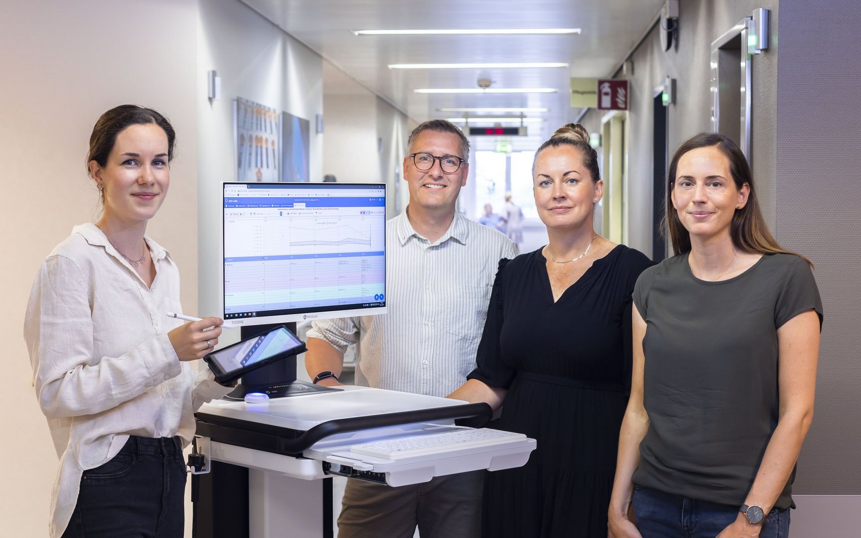 Team mit digitalem Visitenwagen und Patientenakte im Klinikflur.